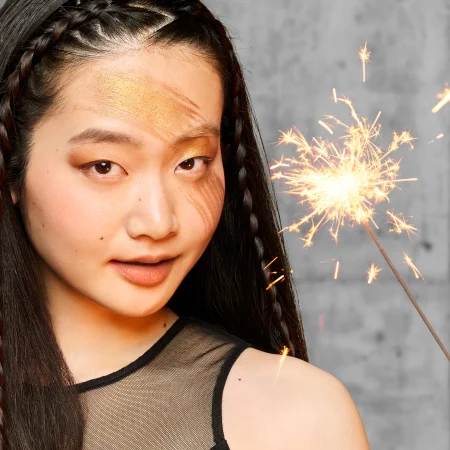 alt="Model with braided hair and bold gold editorial eye makeup holding a sparkling firework against a grey background."
