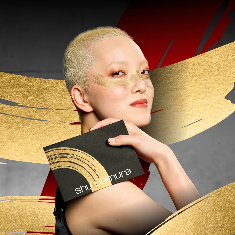 alt="Model with a bleached buzz cut and bold gold editorial makeup holding a black Shu Uemura palette against a dramatic gold and red brushstroke background."