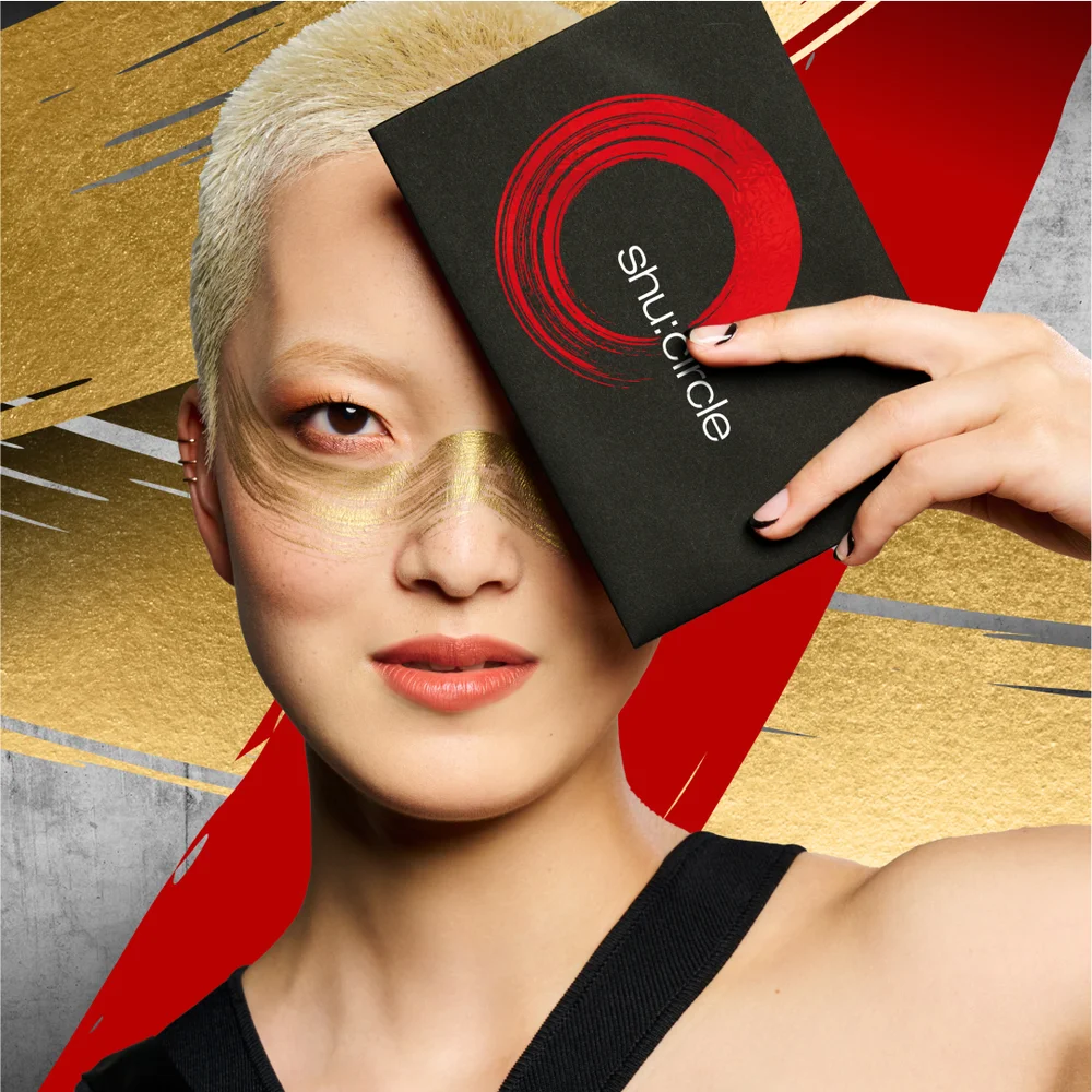 alt="Model with bleached buzz cut and bold gold editorial makeup holding a black Shu Circle loyalty programme box with a red brushstroke logo over one eye, against a gold and red background."